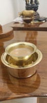 A brass tumbler filled with tea is placed on a polished wooden surface. In the background, there is a decorative statue and some out-of-focus items.