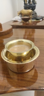 A brass tumbler filled with tea is placed on a polished wooden surface. In the background, there is a decorative statue and some out-of-focus items.