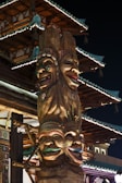 A tall, intricately carved totem pole with four colorful, expressive faces stacked vertically. Each face has unique decorations and vibrant colors, set against a traditional Asian architectural structure with ornate details and illuminated eaves. The scene is captured at night, highlighting the contrast between the warm light on the totem and the dark sky.