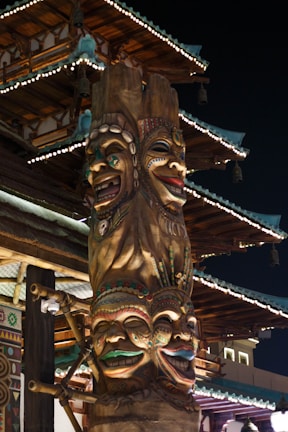 A tall, intricately carved totem pole with four colorful, expressive faces stacked vertically. Each face has unique decorations and vibrant colors, set against a traditional Asian architectural structure with ornate details and illuminated eaves. The scene is captured at night, highlighting the contrast between the warm light on the totem and the dark sky.