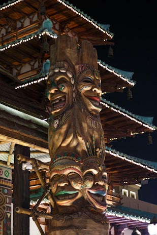 A tall, intricately carved totem pole with four colorful, expressive faces stacked vertically. Each face has unique decorations and vibrant colors, set against a traditional Asian architectural structure with ornate details and illuminated eaves. The scene is captured at night, highlighting the contrast between the warm light on the totem and the dark sky.