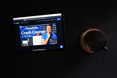 An iPad displaying a website with a blue-themed crash course advertisement, featuring a person in a blue shirt prominently. Beside the iPad lies a dark-colored coffee mug on a wooden coaster, all against a black background.