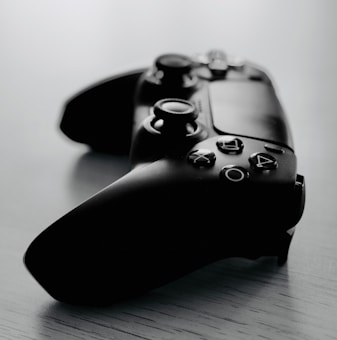 A black gaming controller rests on a pale wooden surface, showing a close-up of its buttons and joysticks. The background is softly blurred, providing a clean and modern aesthetic.