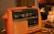 A vintage orange flip clock radio displaying the time as 19:59. The device is slightly dusty and shows signs of wear. It features a manual tuning dial on the side and several wires can be seen nearby on the table.