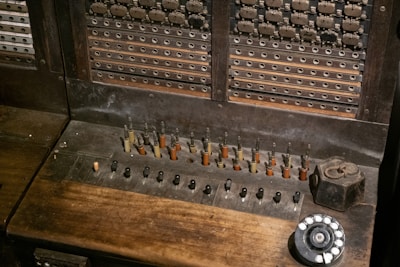 A vintage telephone switchboard featuring numerous metal jacks and plugs, with a rotary dial on the side. The equipment has a worn, wooden surface and metallic components that appear aged and tarnished.