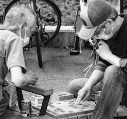 Two people, wearing masks, are deeply engaged in a board game outdoors. They are seated on a bench, with bicycles in the background. Both individuals appear focused and thoughtful, one pointing at the board as they strategize their next move. There are two cans placed beside them.