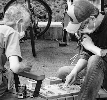 Two people, wearing masks, are deeply engaged in a board game outdoors. They are seated on a bench, with bicycles in the background. Both individuals appear focused and thoughtful, one pointing at the board as they strategize their next move. There are two cans placed beside them.