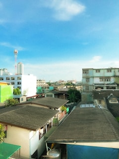 Urban landscape with a clear blue sky overhead. Residential buildings with tiled roofs are visible, surrounded by a mix of greenery and other structures. There is a mix of modern and older architectural styles. A telecom tower is visible in the background.