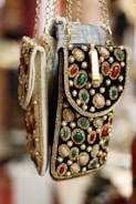 Ornate handbags adorned with colorful gemstones and intricate embroidery hang side by side. The bags feature a combination of red, green, and white stones set against a richly textured fabric. Gold chains and detailing enhance the luxurious appearance.