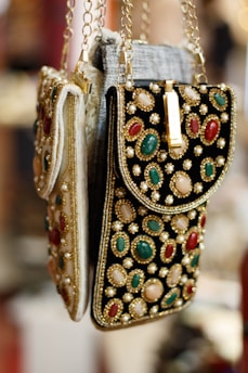 Ornate handbags adorned with colorful gemstones and intricate embroidery hang side by side. The bags feature a combination of red, green, and white stones set against a richly textured fabric. Gold chains and detailing enhance the luxurious appearance.