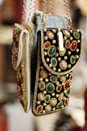 Ornate handbags adorned with colorful gemstones and intricate embroidery hang side by side. The bags feature a combination of red, green, and white stones set against a richly textured fabric. Gold chains and detailing enhance the luxurious appearance.