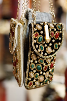 Ornate handbags adorned with colorful gemstones and intricate embroidery hang side by side. The bags feature a combination of red, green, and white stones set against a richly textured fabric. Gold chains and detailing enhance the luxurious appearance.