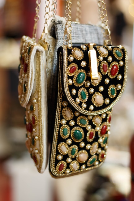 Ornate handbags adorned with colorful gemstones and intricate embroidery hang side by side. The bags feature a combination of red, green, and white stones set against a richly textured fabric. Gold chains and detailing enhance the luxurious appearance.