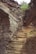A rugged, narrow staircase carved into a rocky terrain leads upwards between steep rock walls. Sparse vegetation, including small shrubs and greenery, clings to the rocky surfaces.