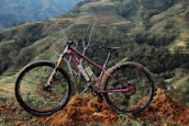 A mountain bike with a dark red frame and black tires is positioned on a mound of dirt. The backdrop consists of terraced hills and lush greenery, suggesting a rural landscape. The bike's tires are muddy, indicating recent off-road use.