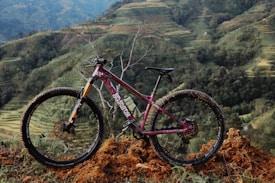 A mountain bike with a dark red frame and black tires is positioned on a mound of dirt. The backdrop consists of terraced hills and lush greenery, suggesting a rural landscape. The bike's tires are muddy, indicating recent off-road use.