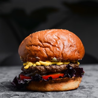 A delicious-looking hamburger with a golden brown bun sits on a grey surface. The burger features layers of grilled beef patty, melted cheese, fresh tomato slices, and leafy lettuce. The arrangement is visually appealing, suggesting a freshly prepared meal.