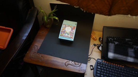 A wooden desk is organized with a black laptop, a colorful book placed centrally on a black mat, a patterned scarf, a wired keyboard with orange keys, and a plant in a glass jar. An orange box is partially visible on the left.