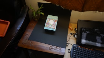 A wooden desk is organized with a black laptop, a colorful book placed centrally on a black mat, a patterned scarf, a wired keyboard with orange keys, and a plant in a glass jar. An orange box is partially visible on the left.