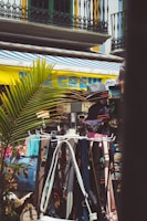 A vibrant outdoor market scene features clothes and accessories on display, including hats, bags, and belts. The bright yellow and blue of an awning contrasts with the ironwork and balconies of nearby buildings. Palm leaves add a tropical element to the setting.