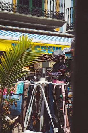 A vibrant outdoor market scene features clothes and accessories on display, including hats, bags, and belts. The bright yellow and blue of an awning contrasts with the ironwork and balconies of nearby buildings. Palm leaves add a tropical element to the setting.