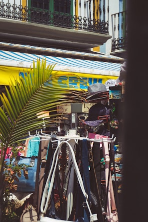 A vibrant outdoor market scene features clothes and accessories on display, including hats, bags, and belts. The bright yellow and blue of an awning contrasts with the ironwork and balconies of nearby buildings. Palm leaves add a tropical element to the setting.