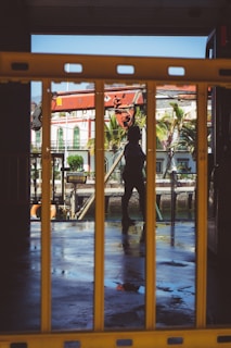 A silhouette of a person is visible through yellow barriers, with the background showing tropical buildings, palm trees, and a partially visible boat or ship. The ground appears wet, reflecting the surrounding environment.