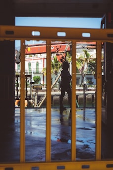 A silhouette of a person is visible through yellow barriers, with the background showing tropical buildings, palm trees, and a partially visible boat or ship. The ground appears wet, reflecting the surrounding environment.