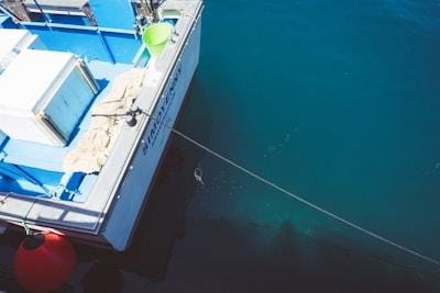 A docked boat with part of its deck visible, featuring a blue color scheme. There is a green bucket and what appears to be a cooler on the deck. The boat is moored with a rope extending into the water, which is a deep, clear blue. A large red buoy is attached to the side of the boat.