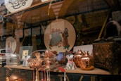 A shop window displays a variety of decorative and crafted items. There are copper pots with a shiny finish, ceramic mugs with floral patterns, and a plate featuring a traditional art design depicting people in colorful attire. The background shows more decorative objects and possibly textiles, adding to the artisanal and cultural theme.