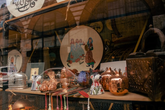 A shop window displays a variety of decorative and crafted items. There are copper pots with a shiny finish, ceramic mugs with floral patterns, and a plate featuring a traditional art design depicting people in colorful attire. The background shows more decorative objects and possibly textiles, adding to the artisanal and cultural theme.