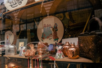 A shop window displays a variety of decorative and crafted items. There are copper pots with a shiny finish, ceramic mugs with floral patterns, and a plate featuring a traditional art design depicting people in colorful attire. The background shows more decorative objects and possibly textiles, adding to the artisanal and cultural theme.