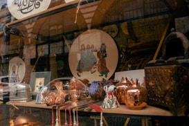 A shop window displays a variety of decorative and crafted items. There are copper pots with a shiny finish, ceramic mugs with floral patterns, and a plate featuring a traditional art design depicting people in colorful attire. The background shows more decorative objects and possibly textiles, adding to the artisanal and cultural theme.