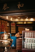 A person wearing a face mask is sitting behind a counter in a traditional herbal medicine store. The store has an ornate interior with gold and red decorations. There is a large silver ceremonial urn to the left and a refrigerator with multiple cups of herbal jelly to the right. The background features shelves with various items and Chinese calligraphy.