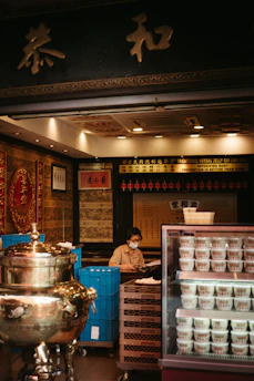 A person wearing a face mask is sitting behind a counter in a traditional herbal medicine store. The store has an ornate interior with gold and red decorations. There is a large silver ceremonial urn to the left and a refrigerator with multiple cups of herbal jelly to the right. The background features shelves with various items and Chinese calligraphy.
