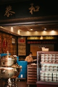 A person wearing a face mask is sitting behind a counter in a traditional herbal medicine store. The store has an ornate interior with gold and red decorations. There is a large silver ceremonial urn to the left and a refrigerator with multiple cups of herbal jelly to the right. The background features shelves with various items and Chinese calligraphy.
