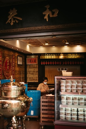 A person wearing a face mask is sitting behind a counter in a traditional herbal medicine store. The store has an ornate interior with gold and red decorations. There is a large silver ceremonial urn to the left and a refrigerator with multiple cups of herbal jelly to the right. The background features shelves with various items and Chinese calligraphy.