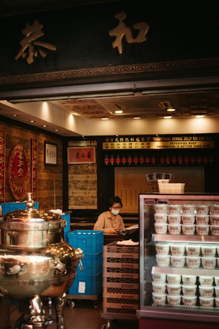 A person wearing a face mask is sitting behind a counter in a traditional herbal medicine store. The store has an ornate interior with gold and red decorations. There is a large silver ceremonial urn to the left and a refrigerator with multiple cups of herbal jelly to the right. The background features shelves with various items and Chinese calligraphy.