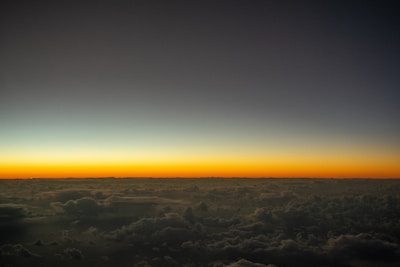 A breathtaking view of a horizon at sunset or sunrise with a gradient of colors transitioning from dark blue to light orange. Below the horizon, fluffy clouds spread out, creating a serene and expansive atmosphere.