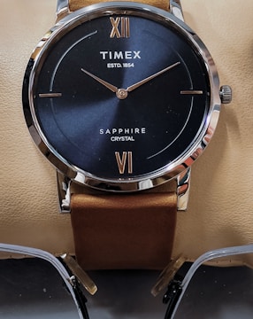 A close-up view of a wristwatch with a navy blue face and silver-colored rim. The watch features Roman numerals XII and VI, and the brand name 'Timex' along with 'Sapphire Crystal' on the dial. The watch has a brown leather strap and is placed on a light beige surface. Below the watch, a pair of glasses with a metallic frame is partially visible.