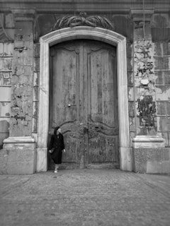 A person dressed in dark clothing leans against an old, ornate wooden door set in a weathered stone façade. The door is large, with intricate carvings and signs of age, surrounded by a worn, textured wall. The atmosphere is historic and possibly melancholic.