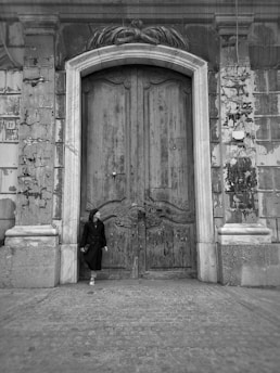A person dressed in dark clothing leans against an old, ornate wooden door set in a weathered stone façade. The door is large, with intricate carvings and signs of age, surrounded by a worn, textured wall. The atmosphere is historic and possibly melancholic.