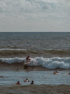 A surfer rides a wave in the ocean while several people swim nearby. The scene is set under a cloudy sky, with the ocean's waves capturing the light, creating a dynamic and adventurous atmosphere.