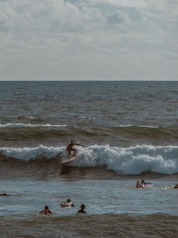 A surfer rides a wave in the ocean while several people swim nearby. The scene is set under a cloudy sky, with the ocean's waves capturing the light, creating a dynamic and adventurous atmosphere.