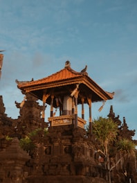 An intricately designed traditional Balinese temple structure with ornate carvings and a bright orange-tiled roof stands prominently against a blue sky. Surrounding the temple are dark stone walls adorned with green plants and unique architectural details.