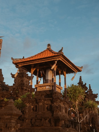 An intricately designed traditional Balinese temple structure with ornate carvings and a bright orange-tiled roof stands prominently against a blue sky. Surrounding the temple are dark stone walls adorned with green plants and unique architectural details.