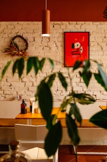 A cozy cafe interior features a white brick wall adorned with a vibrant red artwork. A wreath made of dried flowers hangs on the wall beside the painting. A wooden table is set with condiments and napkins, while a modern brown pendant light hangs overhead. Green plant leaves partially cover the foreground, adding a natural element.