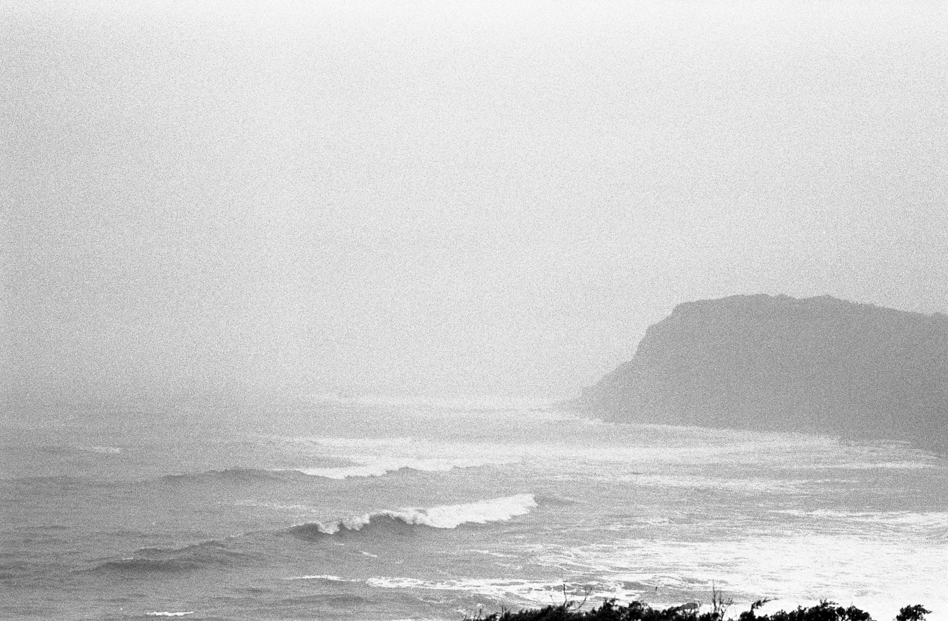 A dramatic black and white image of a rocky beach shoreline with crashing waves and distant cliffs.