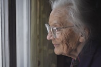 an old lady with glasses looks out of a window. She has a slightl smile on her face.