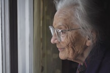 an old lady with glasses looks out of a window. She has a slightl smile on her face.